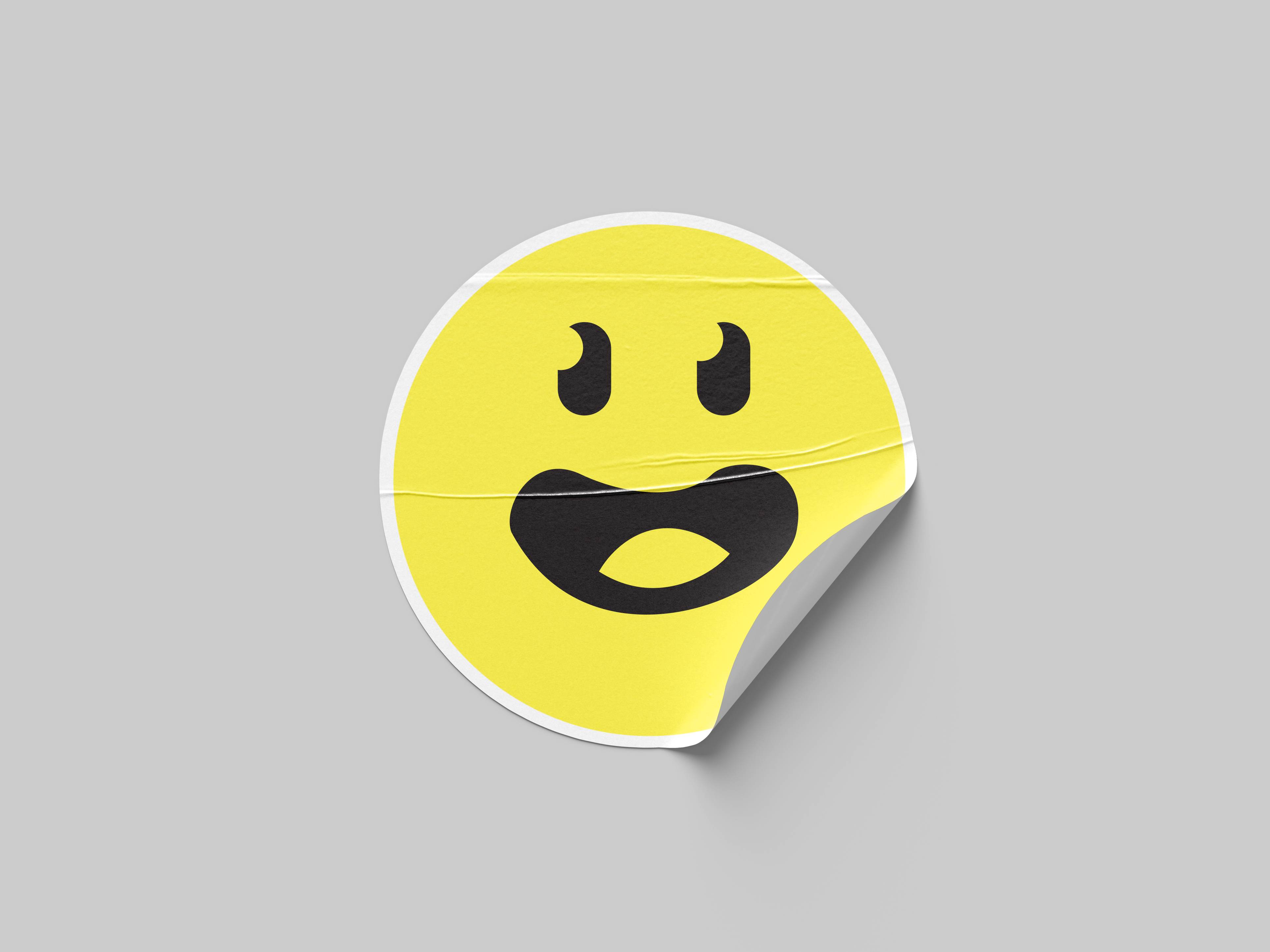 A round yellow Sculoo face sticker with a big open-mouthed grin on pale grey, its bottom right edge peeling up