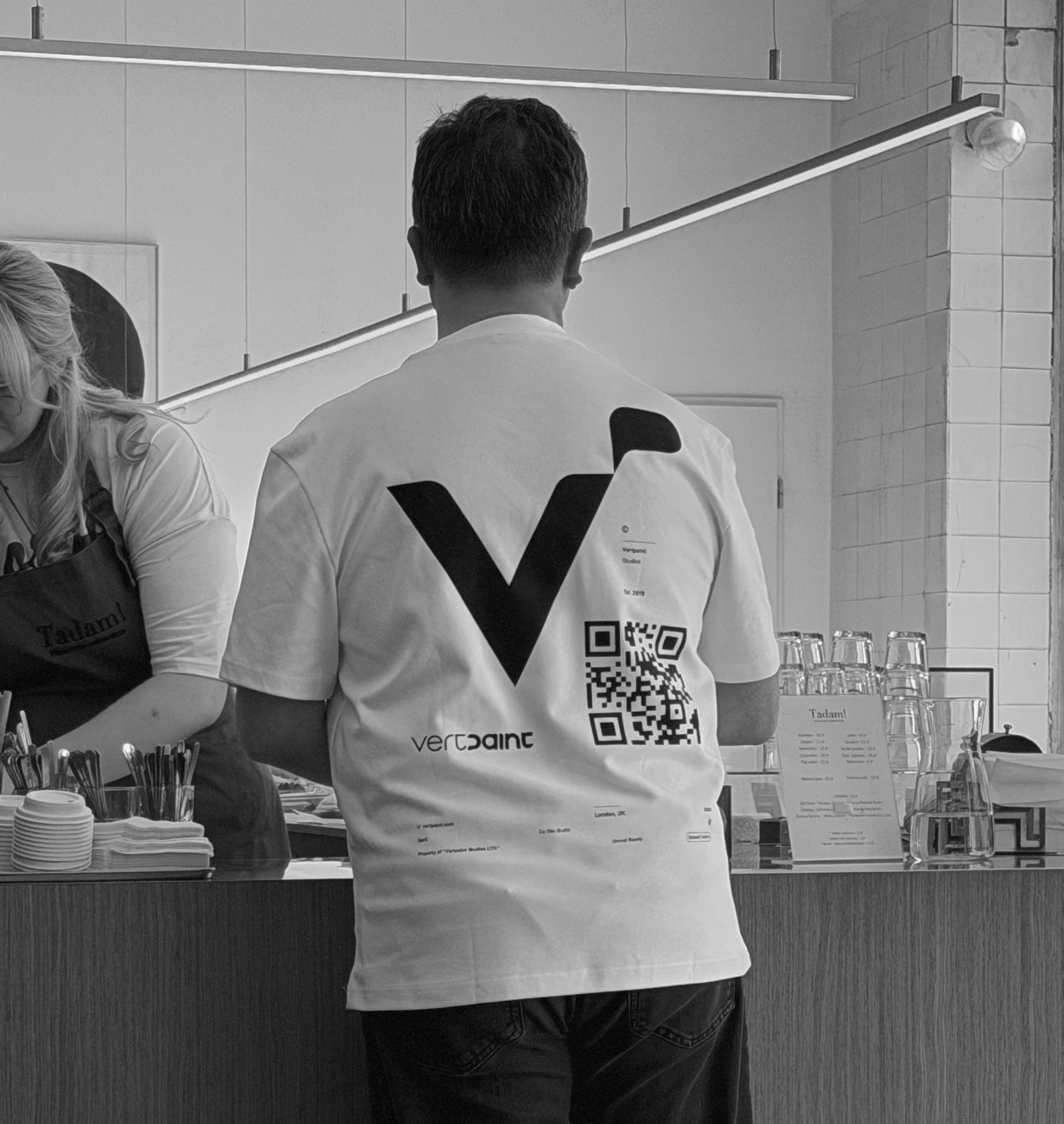 A man at a café counter wearing a white Vertpaint T-shirt with a large V on the back